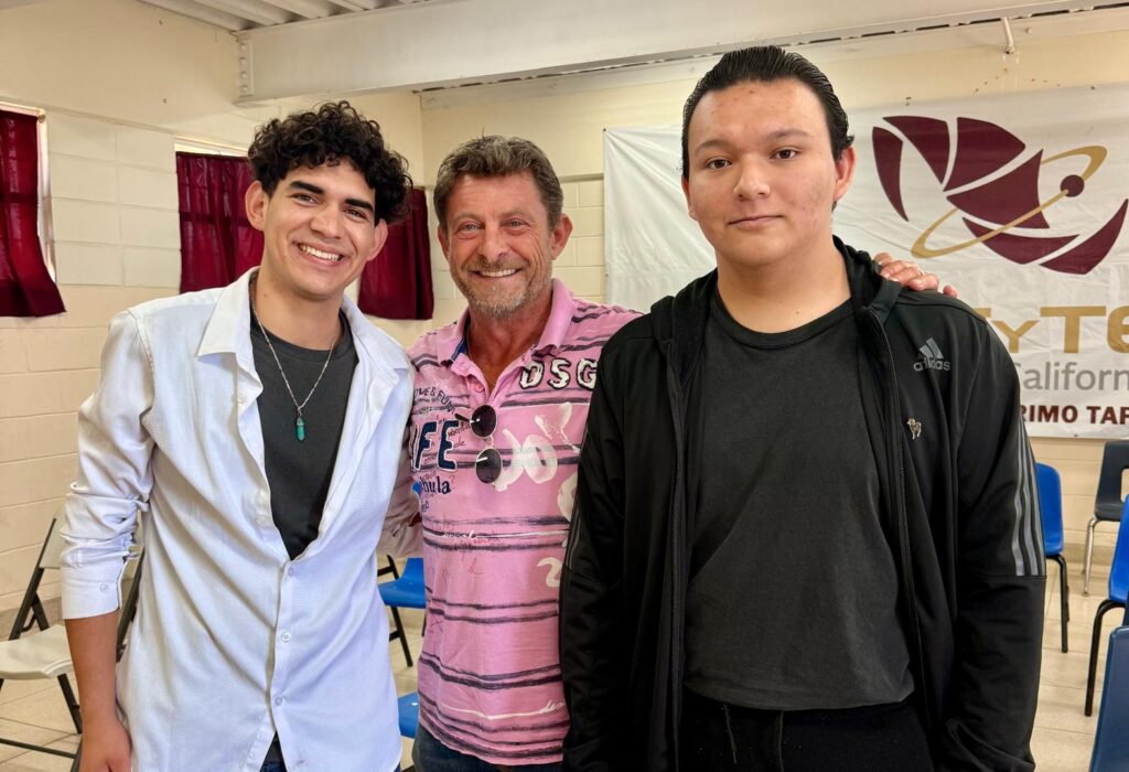 Oswaldo and Christian stand with Baja Angels of the Sea founder Bruno, smiling together inside the high school auditorium following the Rotary Scholars scholarship presentation in Baja California